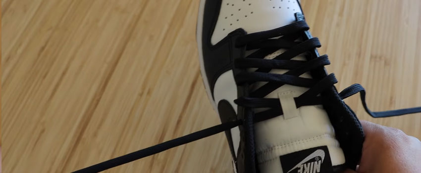 A close-up image of a black and white Nike Dunk sneaker, emphasizing the crisscross lacing style. The sneaker is set against a light wooden floor, with a hand holding one of the laces, highlighting the shoe's design details and brand logo.