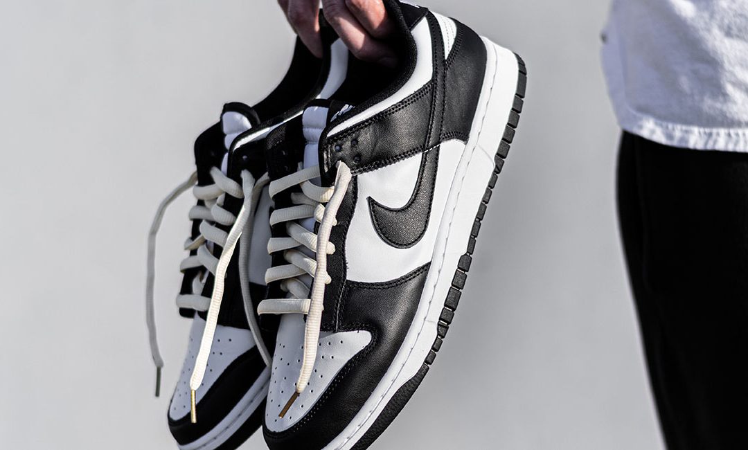 A person holding a pair of black and white Nike sneakers against a neutral backdrop. The image captures the contrasting laces, detailed stitching, and the iconic Nike swoosh. The photo composition includes a partial view of the individual's clothing, suggesting a casual style.
